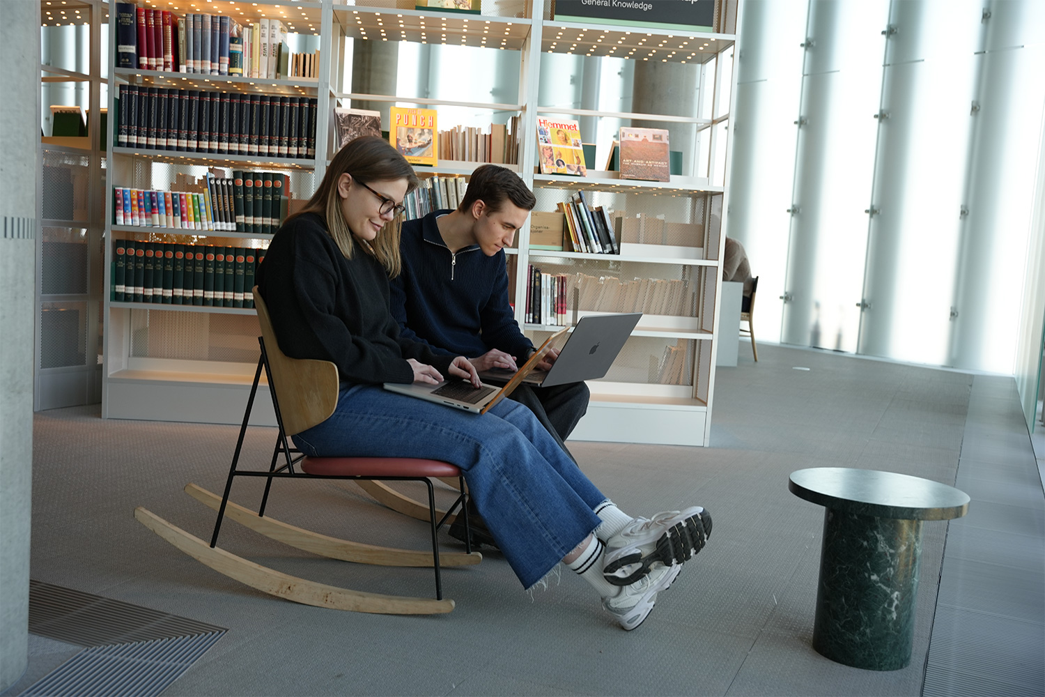 Bildet viser to studenter som sitter i hver sin stol på et bibliotek. De leser på hver sin PC-skjerm.