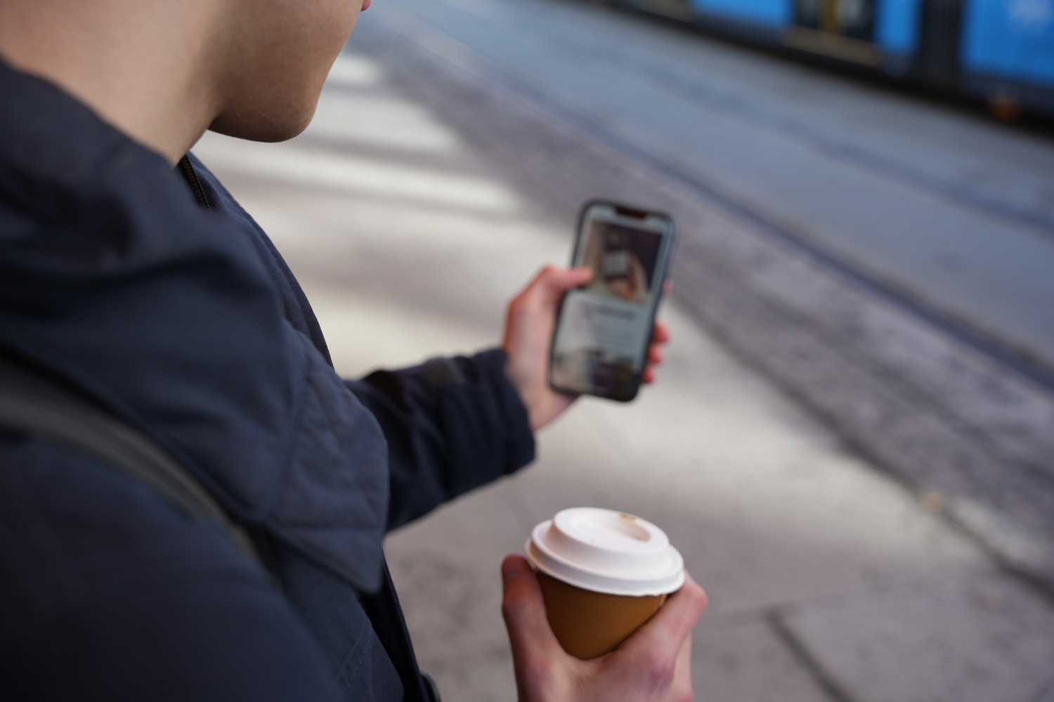 Bildet viser en mann som holder en mobil i hånden mens han venter på trikken. På mobilskjermen vises kunne.no